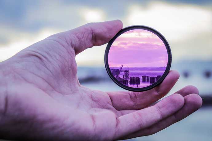 A hand holding a tinted lens that changes the color of the sea beyond from blue to purple